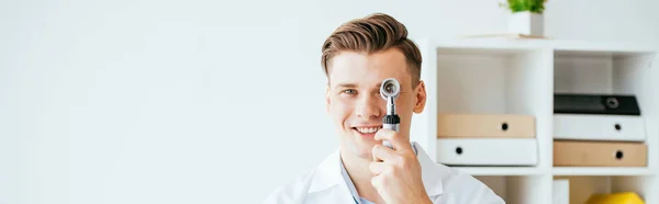 Plano panorámico de alegre dermatólogo en bata blanca sosteniendo dermatoscopio y sonriendo en la clínica - foto de stock