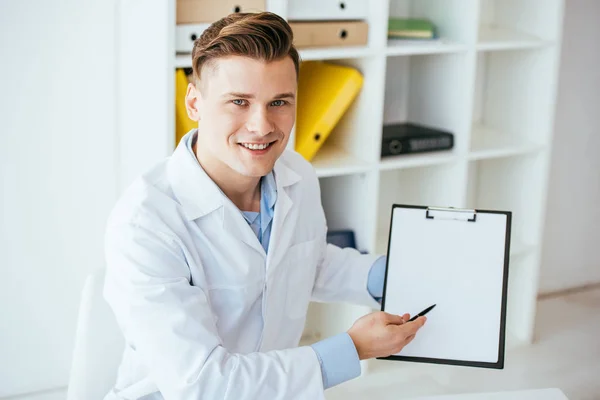 Cheerful doctor in white coat holding pen and blank clipboard — Stock Photo