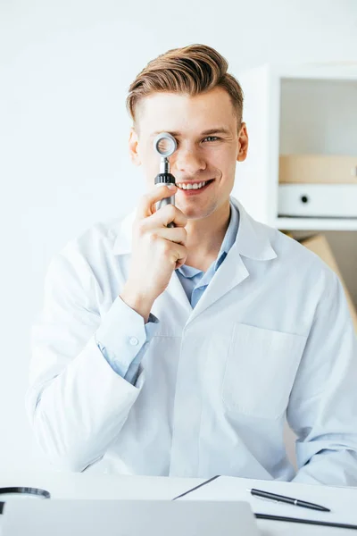 Guapo dermatólogo en blanco abrigo celebración dermatoscopio y sonriendo en clínica - foto de stock