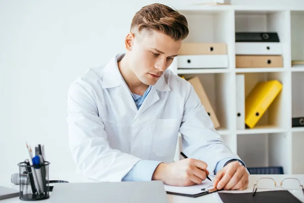 Médecin sérieux et beau en manteau blanc écriture diagnostic sur presse-papiers — Photo de stock