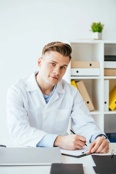 Handsome doctor in white coat holding pen near clipboard — Stock Photo