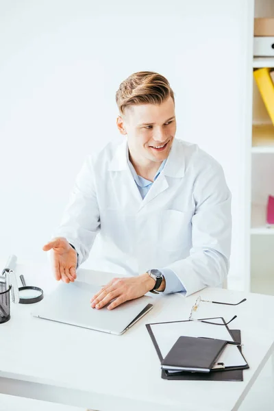 Médecin joyeux en manteau blanc gesticulant près d'un ordinateur portable à la clinique — Photo de stock