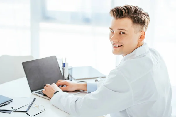 Médico alegre sonriendo mientras usa el ordenador portátil con pantalla en blanco - foto de stock