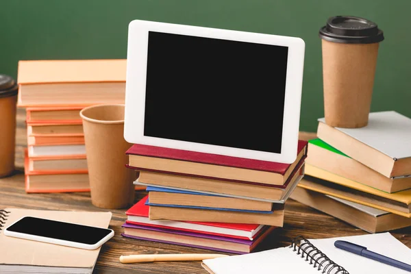 Libri, quaderni, penne, bicchieri usa e getta, smartphone e tablet digitale con schermo bianco su superficie in legno isolata su verde — Foto stock