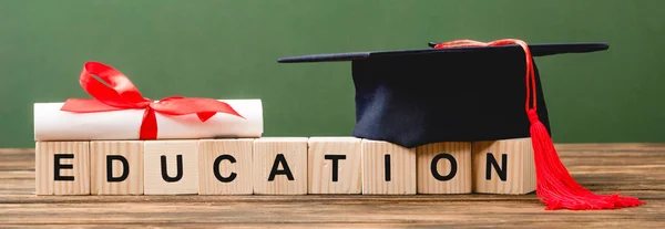 Panoramic shot of wooden blocks with letters, diploma and academic cap on wooden surface isolated on green — Stock Photo