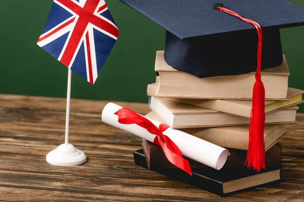 Libros, gorra académica, diploma y bandera británica en superficie de madera aislada en verde - foto de stock
