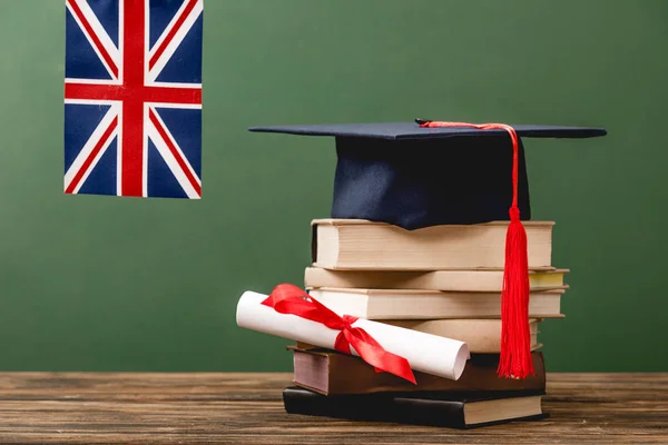 Libros, gorra académica, diploma y bandera británica en superficie de madera aislada en verde - foto de stock
