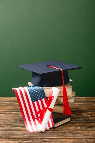Libros, gorra académica, diploma y bandera americana sobre superficie de madera aislada en verde - foto de stock