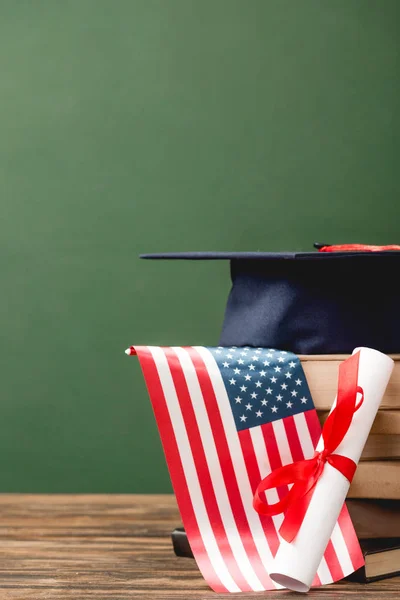Libros, gorra académica, diploma y bandera americana sobre superficie de madera aislada en verde - foto de stock