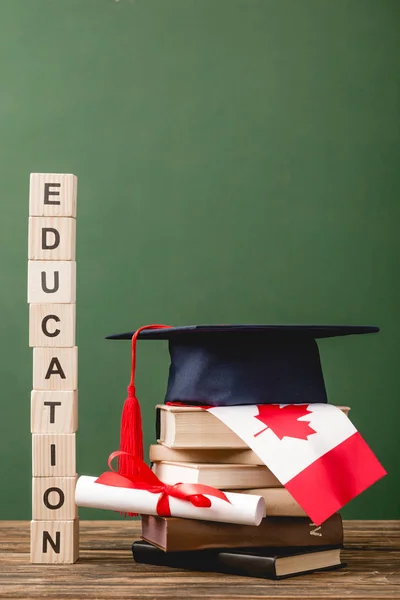 Bloques de madera con letras, gorra académica, libros, diploma y bandera canadiense en la superficie de madera aislada en verde - foto de stock