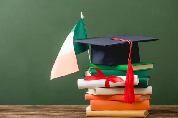 Livros, diploma, boné acadêmico e bandeira irlandesa na superfície de madeira isolada em verde — Fotografia de Stock