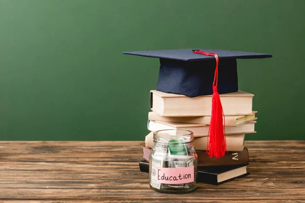 Libros, gorra académica y alcancía en superficie de madera aislada en verde - foto de stock