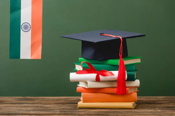 Libros, diploma, gorra académica y bandera india en superficie de madera aislada en verde - foto de stock