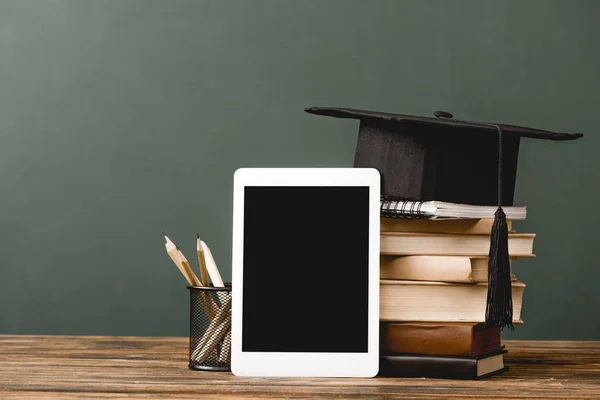 Libros, cuaderno, gorra académica y tableta digital con pantalla en blanco sobre superficie de madera aislada en gris - foto de stock