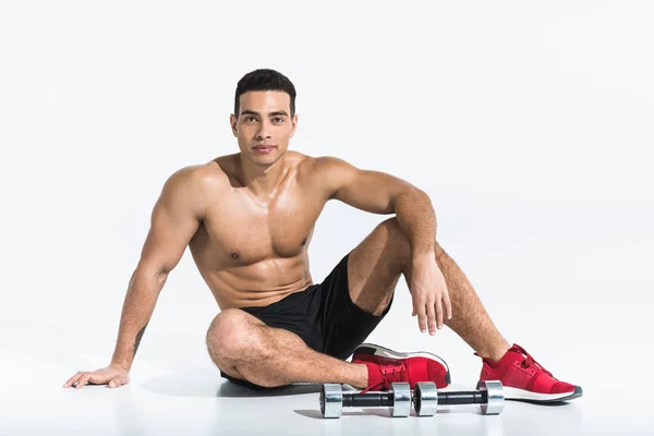 Handsome sportive mixed race man sitting on floor near dumbbells on white — Stock Photo