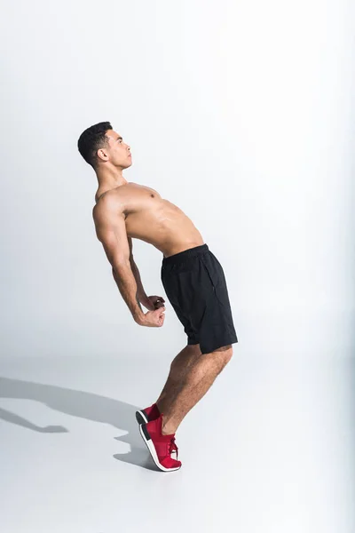 Side view of sportive, handsome mixed race man in black shorts and red sneakers warming up on white — Stock Photo