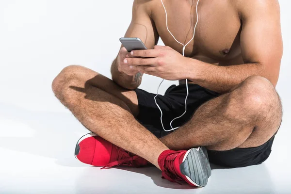 Обрезанный вид спортивного человека со смартфоном, сидящего на полу со скрещенными ногами на белом — стоковое фото