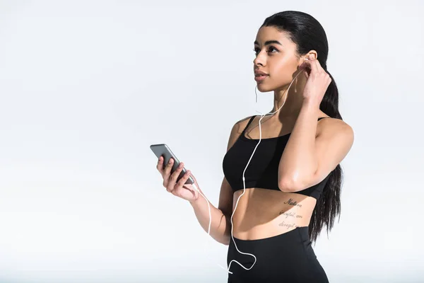 Atractiva chica afroamericana deportiva sosteniendo teléfono inteligente y escuchando música en auriculares en blanco - foto de stock