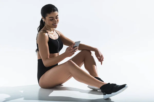 Pretty sportive african american girl sitting on floor, listening music and smiling on white — Stock Photo