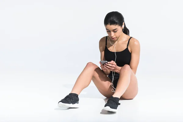 Jolie fille afro-américaine en vêtements de sport assis sur le sol et écouter de la musique dans les écouteurs sur blanc — Photo de stock