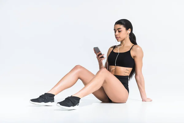 Atractiva chica afroamericana en ropa deportiva sentado en el suelo y escuchar música en los auriculares en blanco - foto de stock