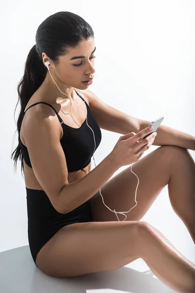 Pretty sportive african american girl with smartphone sitting on floor and listening music in earphones on white — Stock Photo