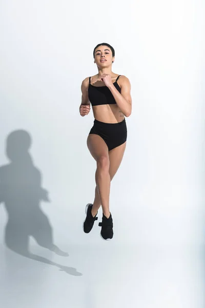 Attraente ragazza africana americana in nero reggiseno sportivo, pantaloncini e scarpe da ginnastica in esecuzione su bianco — Foto stock