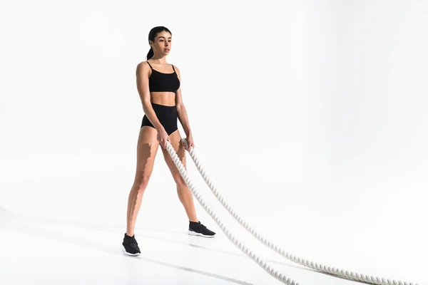 Beautiful young african american woman in black sports bra, short and sneakers holding white ropes on white — Stock Photo