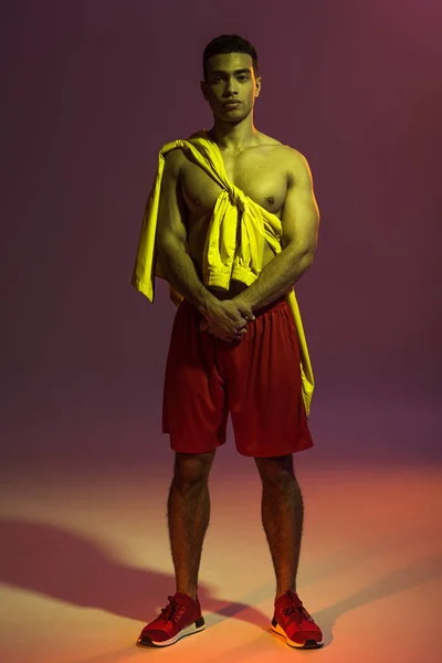 Handsome mixed race man in red shorts, and yellow jacket tied on torso looking at camera on dark background — Stock Photo