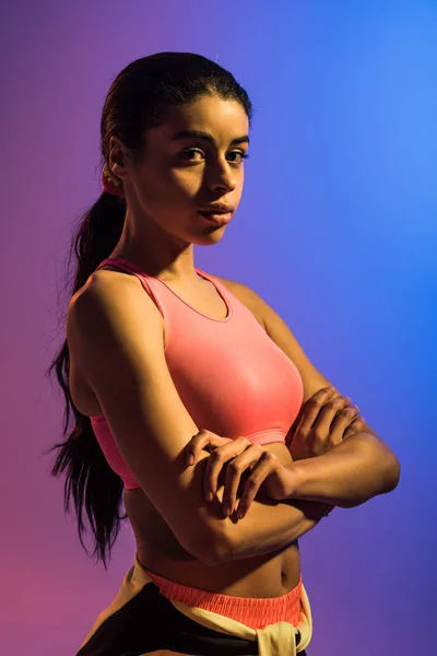 Красива спортивна афроамериканська дівчина позує на камеру з схрещеними руками на фіолетовому і синьому градієнтному фоні — стокове фото
