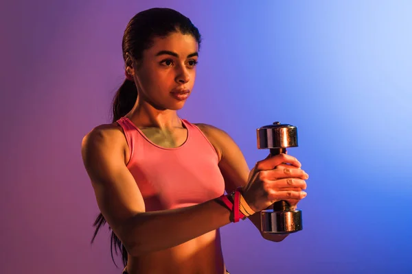 Attractive african american woman in pink sports bra holding dumbbell on purple and blue gradient background — Stock Photo