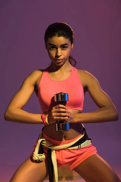 Atletica ragazza afro-americana in possesso di manubri e guardando la fotocamera su sfondo viola — Foto stock