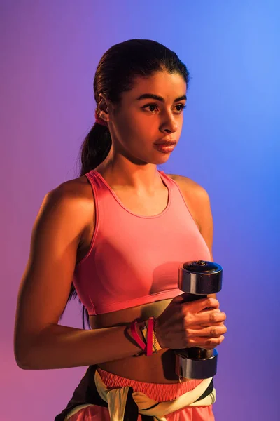 Beautiful african american girl in pink sports bra holding dumbbell on purple and blue gradient background — Stock Photo
