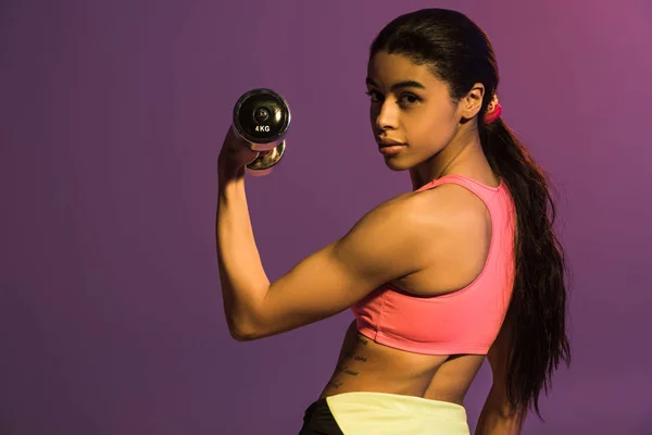 Chica afroamericana bonita en rosa entrenamiento sujetador deportivo con mancuerna y mirando a la cámara aislada en púrpura - foto de stock