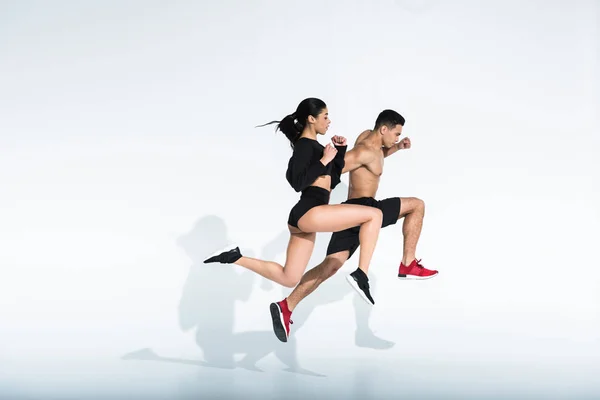 Vista lateral de la mujer y el hombre multicultural deportivo en ropa deportiva y zapatillas de deporte corriendo sobre fondo blanco - foto de stock