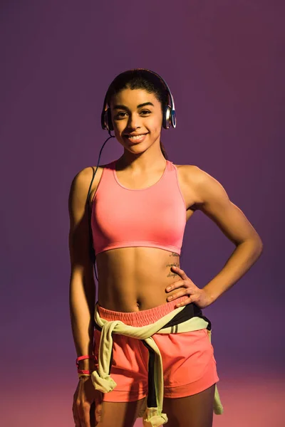 Chica afroamericana deportiva en sujetador deportivo rosa escuchando música en auriculares sobre fondo púrpura - foto de stock