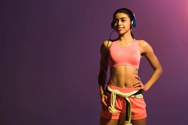 Bela menina africana americana esportiva ouvir música em fones de ouvido e sorrindo no fundo roxo — Fotografia de Stock