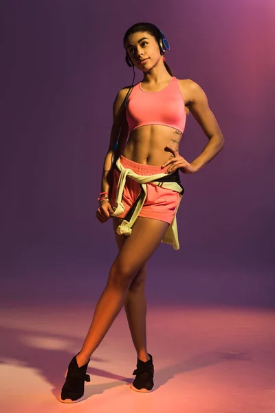 Pretty african american girl in pink sports bra and headphones posing at camera with hand on hip on purple background — Stock Photo