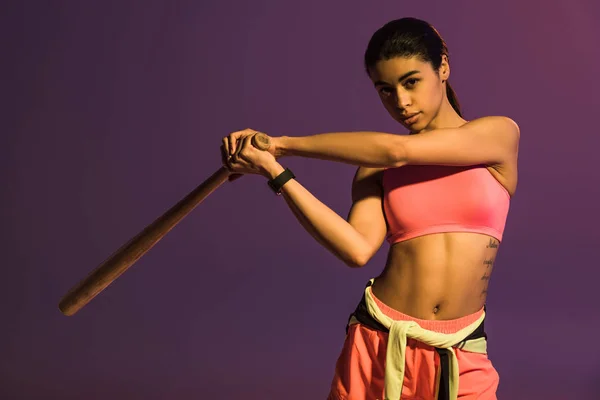 Sportive african american girl in pink sports bra holding baseball bat and looking at camera isolated on purple — Stock Photo