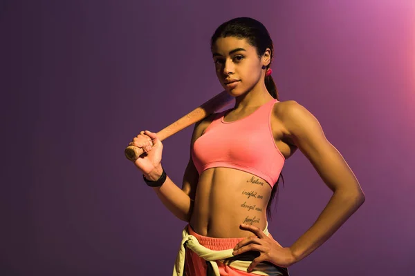 Attractive african american girl with baseball bat posing at camera with hand on hip on purple background — Stock Photo