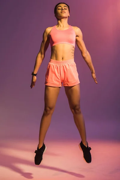 Atractiva chica afroamericana en sujetador deportivo rosa y zapatillas negras saltando con los ojos cerrados sobre fondo púrpura - foto de stock