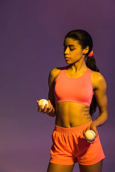 Attractive sportive african american girl in pink sports bra and shorts holding balls on purple background — Stock Photo
