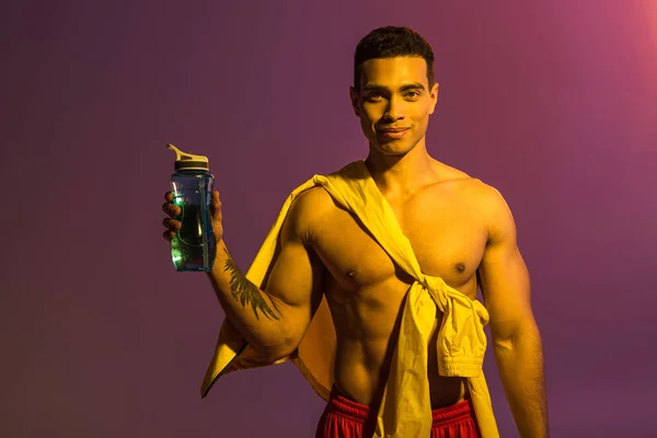 Sonriente guapo mestizo raza hombre con muscular torso celebración deporte botella sobre púrpura fondo - foto de stock