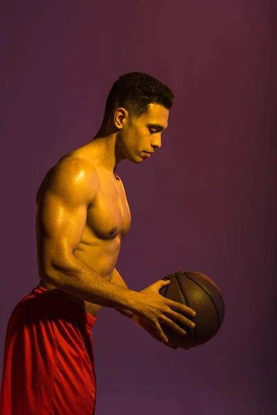 Athletic mixed race man with muscular torso holding brown ball on purple background — Stock Photo