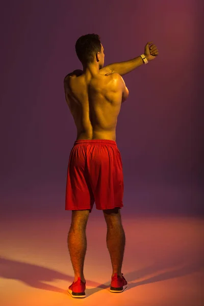 Vista trasera del atleta hombre en pantalones cortos rojos calentándose sobre fondo púrpura - foto de stock