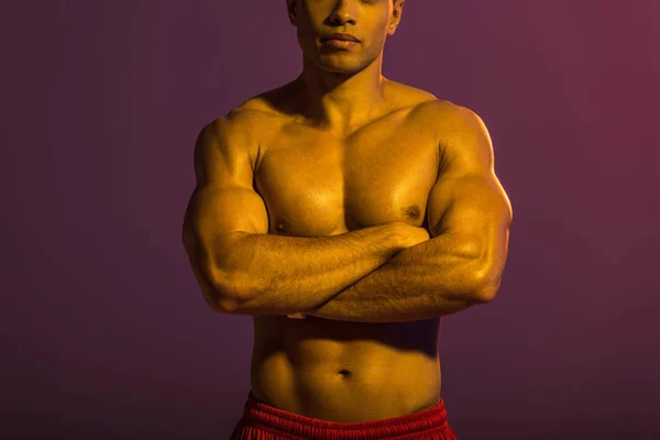 Cropped view of athletic man posing at camera with crossed arms on purple background — Stock Photo