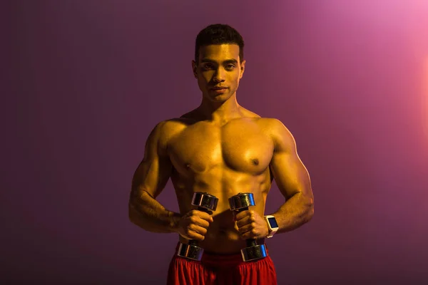 Handsome sportive mixed race man holding dumbbells on purple background — Stock Photo