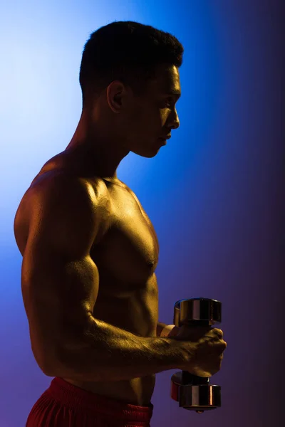 Side view of athletic mixed race man with dumbbells on blue and dark purple gradient background — Stock Photo