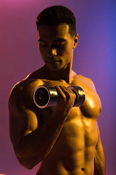 Handsome sportive mixed race man training with dumbbell on purple and blue gradient background — Stock Photo