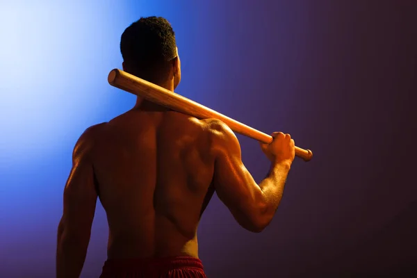 Vista posterior del atlético hombre de raza mixta con bate de béisbol sobre fondo degradado azul y púrpura oscuro — Stock Photo